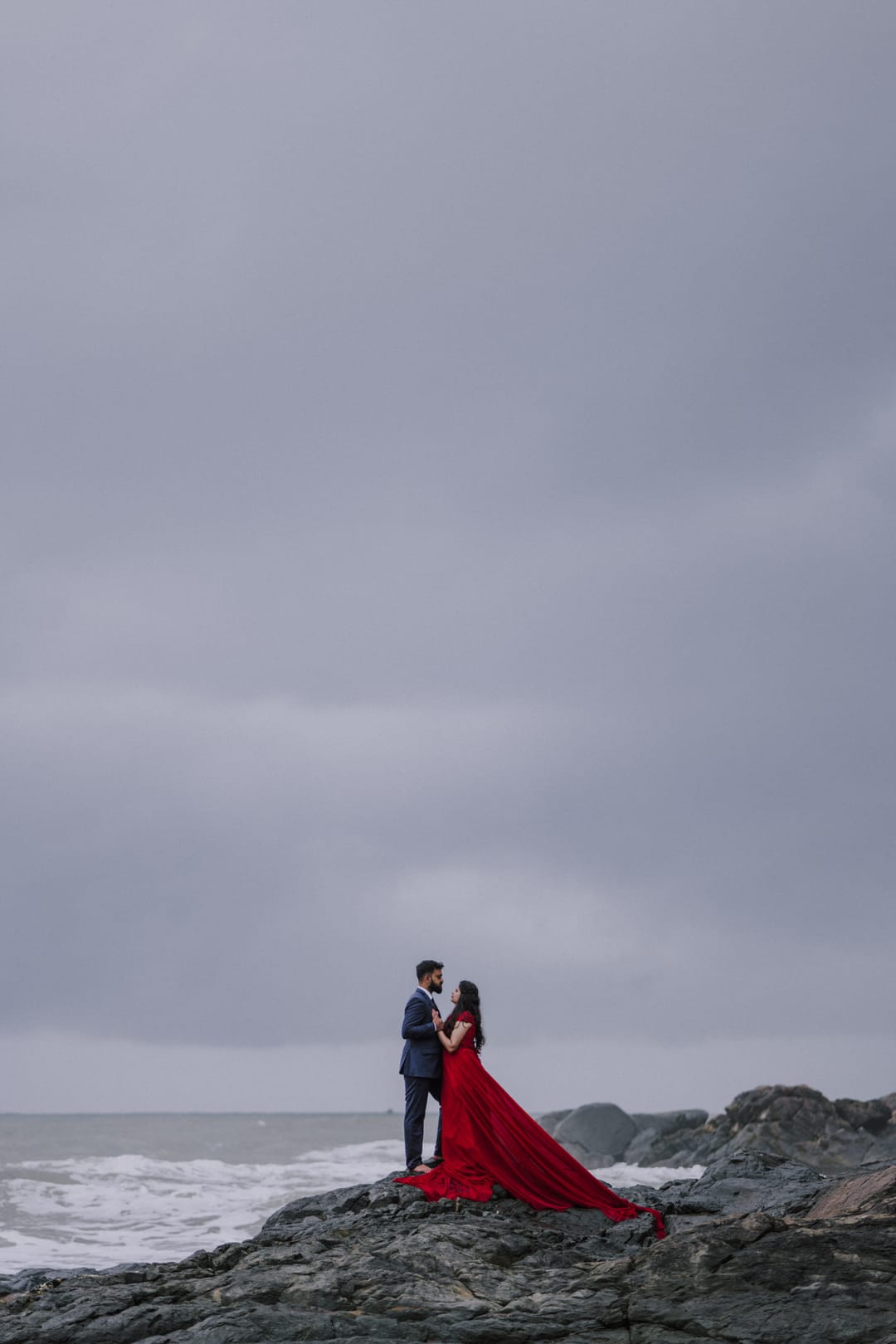 A couple embraces on dark sea rocks under a grey sky, her long red gown trailing over the stones.