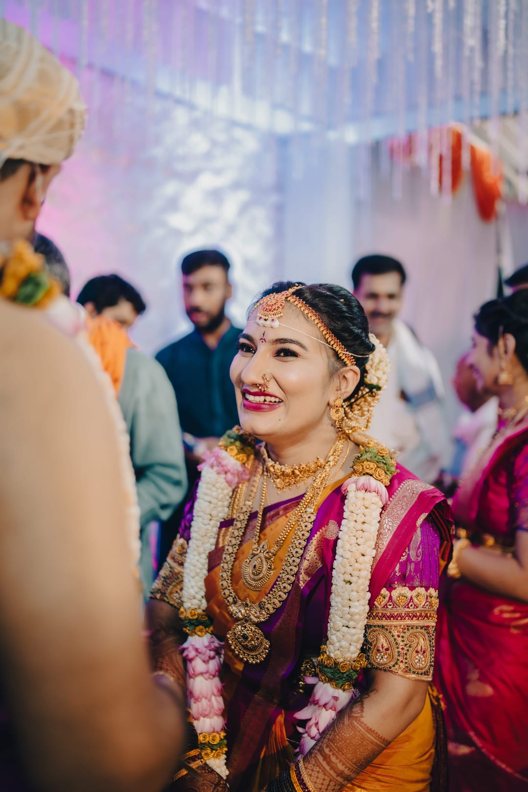 A smiling bride in a pink and gold silk saree and a jasmine garland greets her guests.