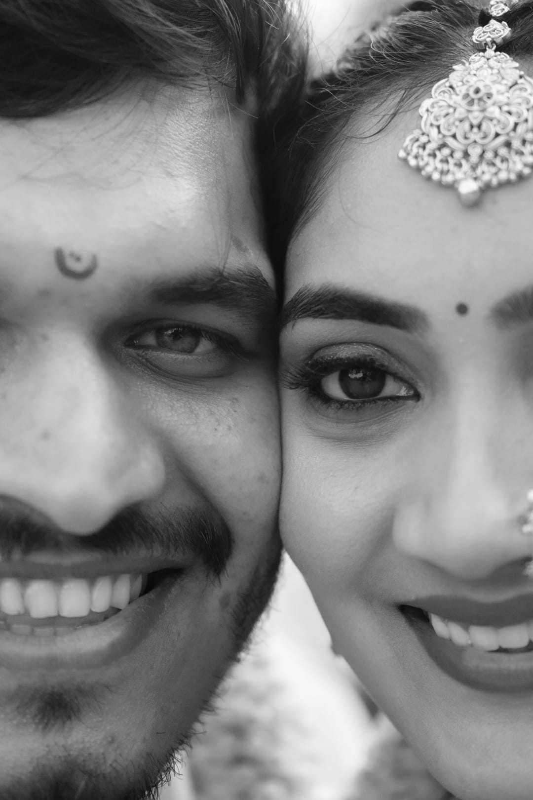 Black-and-white close-up of a smiling couple pressed cheek to cheek, the bride's maang tikka catching the light.