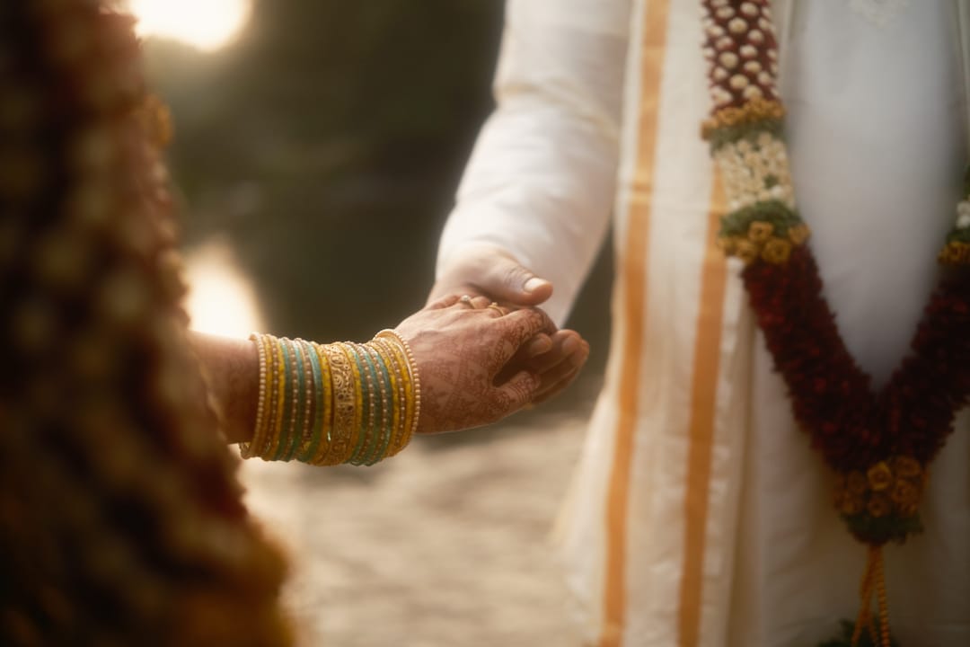 The bride's bangled hand holds the groom's as he stands in a white veshti and a marigold garland, in soft evening light.