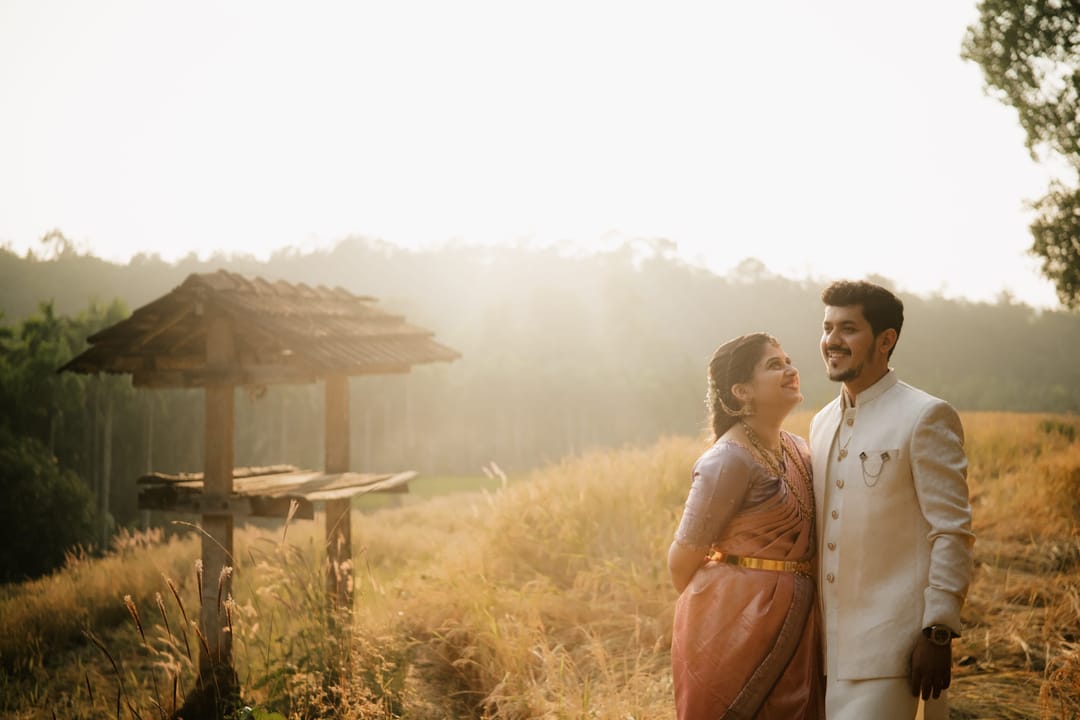 A couple laughs together in golden light on a grassy hillside beside a thatched gazebo.