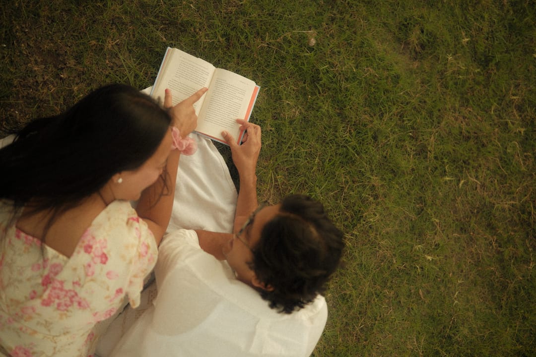 Seen from above, a couple lies on the grass reading a book together, her floral dress spread around them.
