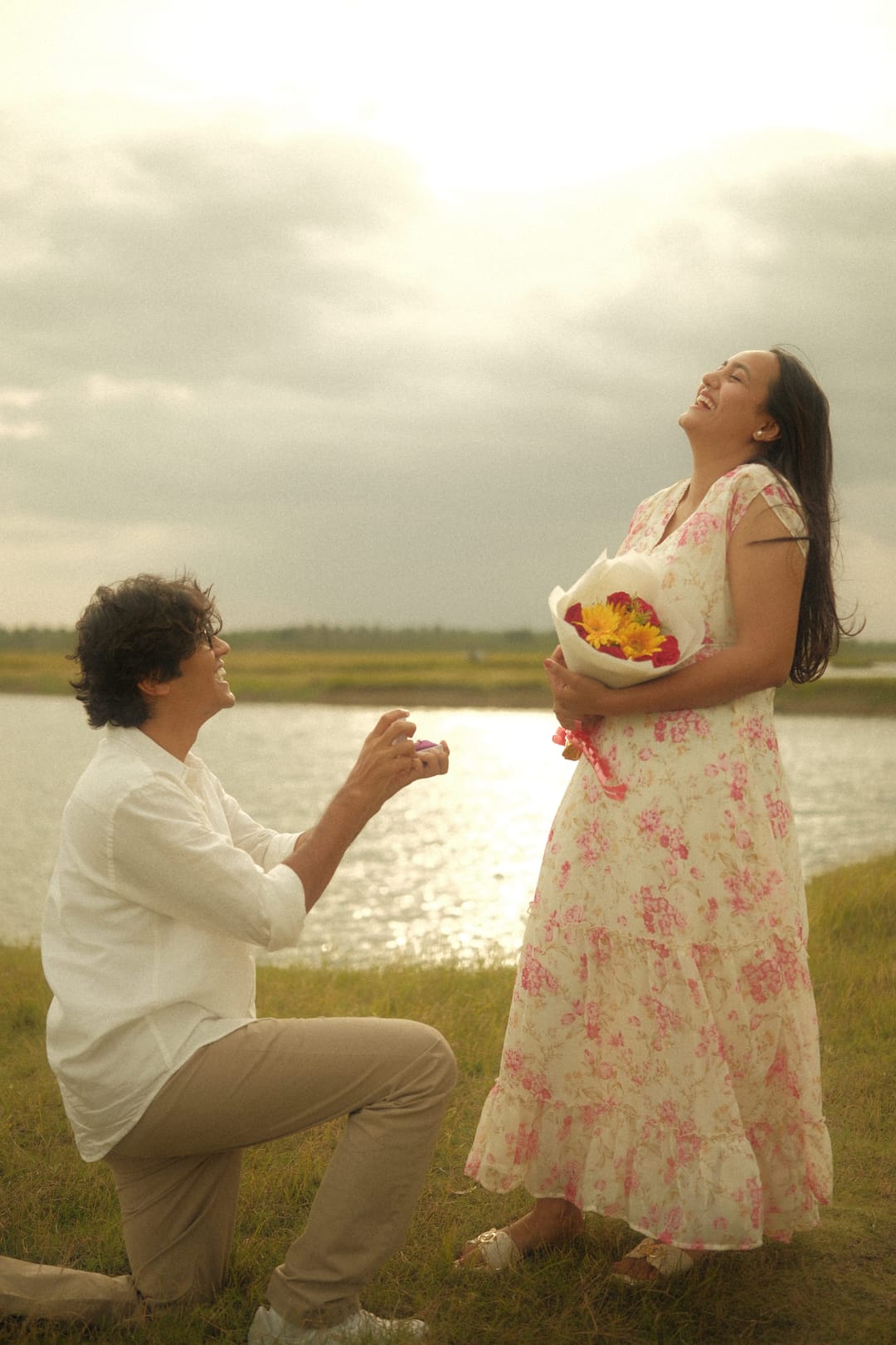 By a sunlit lake, he kneels with a ring as she laughs, holding a bouquet of sunflowers and roses.
