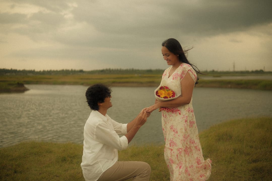 By the water at dusk, he kneels and slips a ring on her finger while she holds a bouquet.