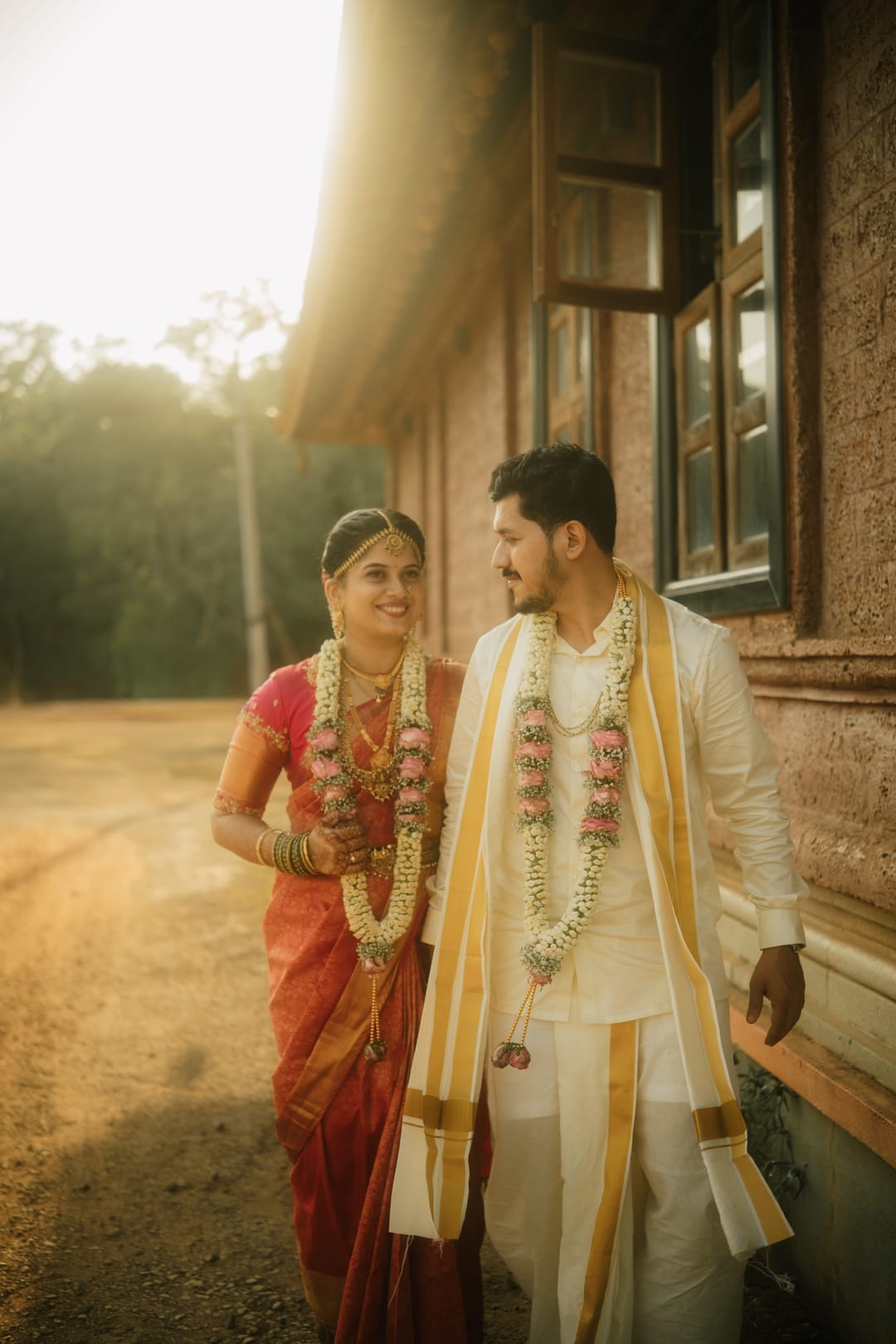 The bride in a red silk saree and the groom in a white veshti walk past an old brick house in late sun, both wearing jasmine garlands.