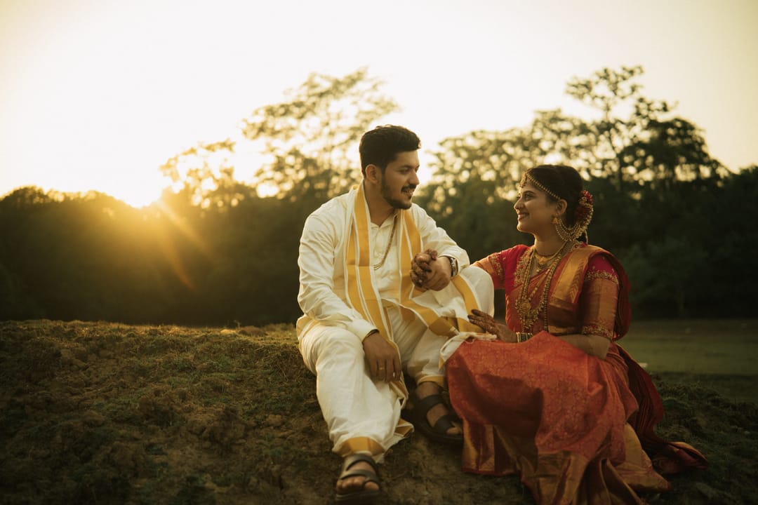 The couple sits on the ground in traditional silks at sunset, sharing a laugh as the sun flares through the trees.