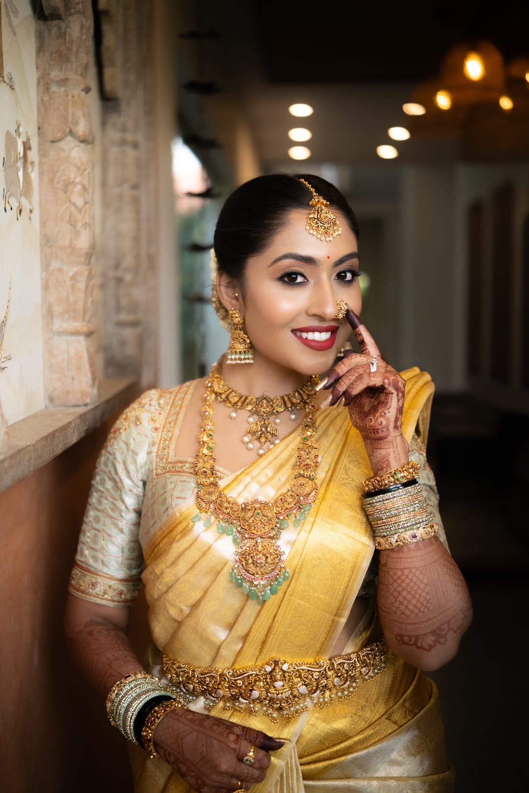 A bride in a golden silk saree and temple jewellery smiles, one hennaed hand raised to her face.