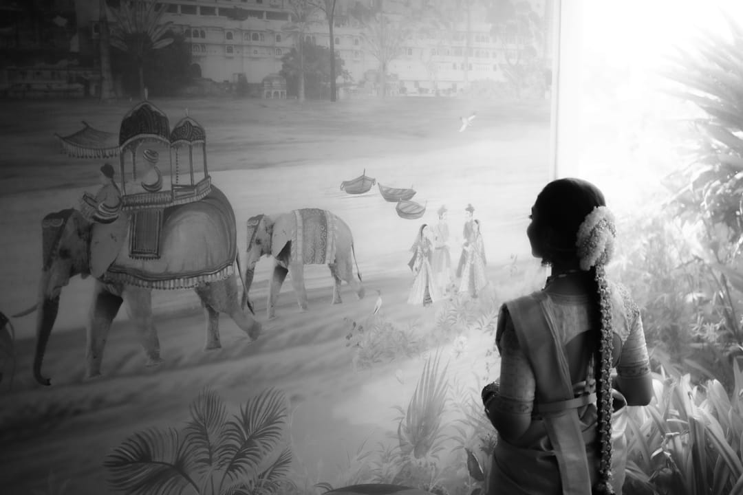 Black-and-white: a bride with a jasmine-wrapped braid looks at a mural of elephants in procession.