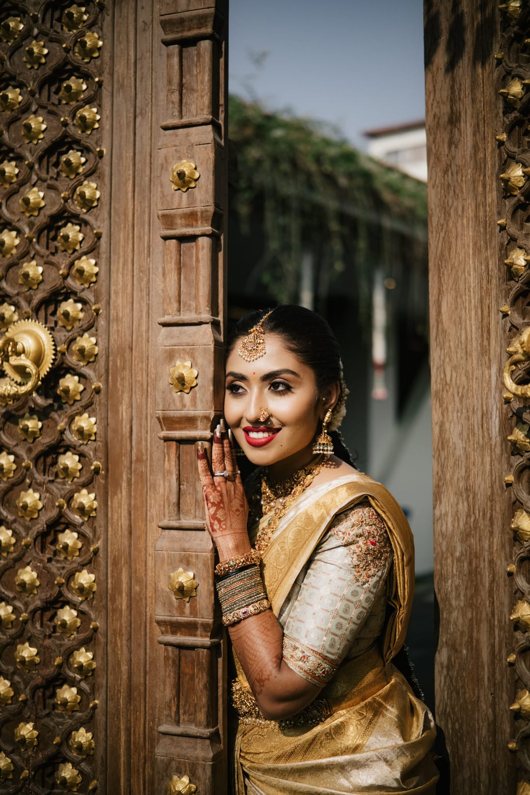 A bride in a gold saree peeks out from behind a carved wooden door, laughing.