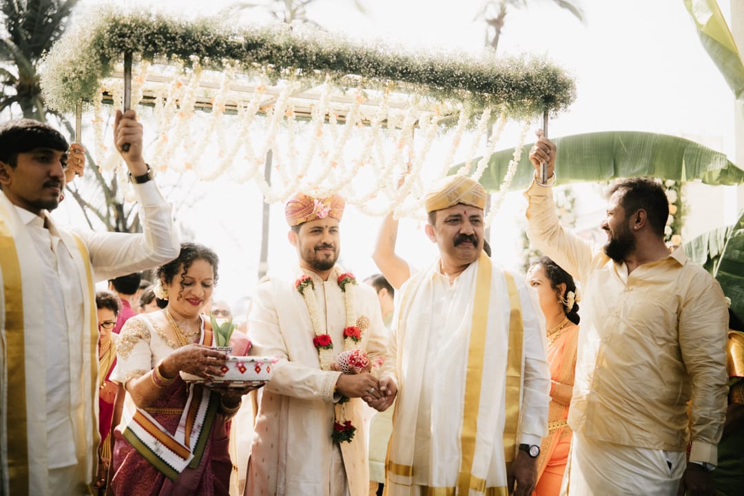 The groom, in a cream sherwani and peta, is led under a jasmine canopy carried by family as his parents hold his hands.