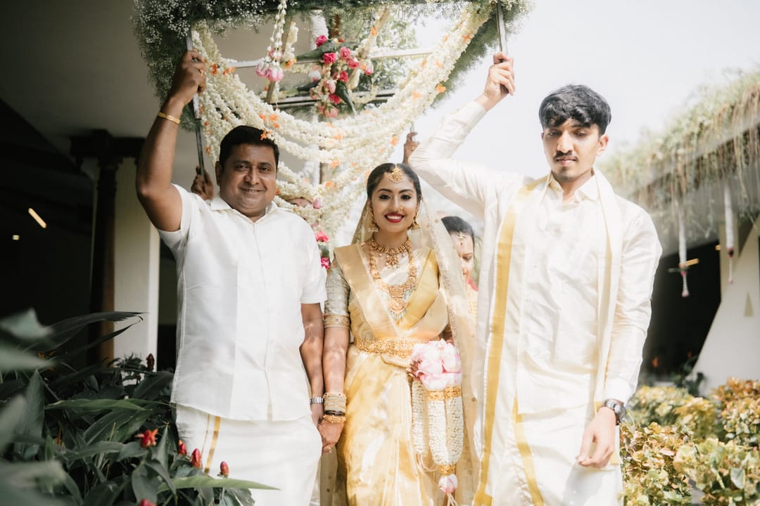 The bride walks in under a flower canopy held up by family, smiling between two relatives.