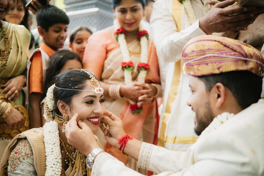 At the ceremony, the groom gently lifts the bride's chin as family gathers close around them.