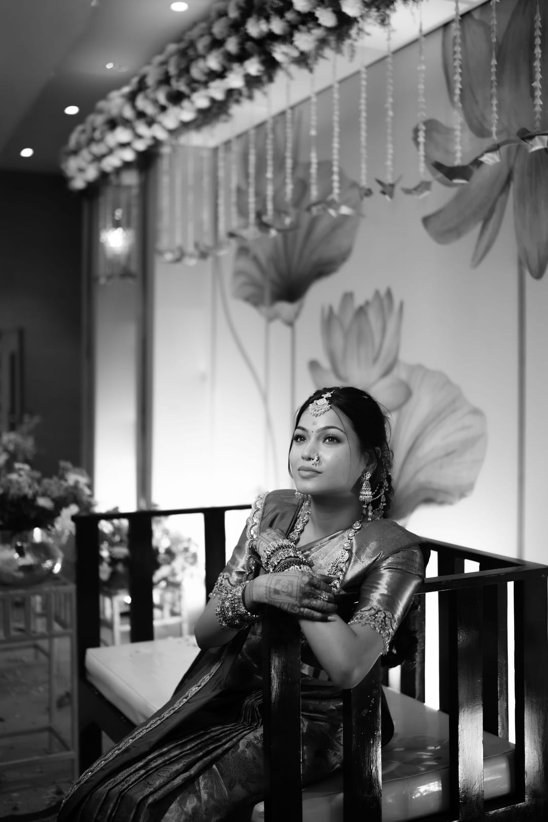 Black-and-white: a bride sits on a carved chair in a silk saree, gazing past painted lotuses.