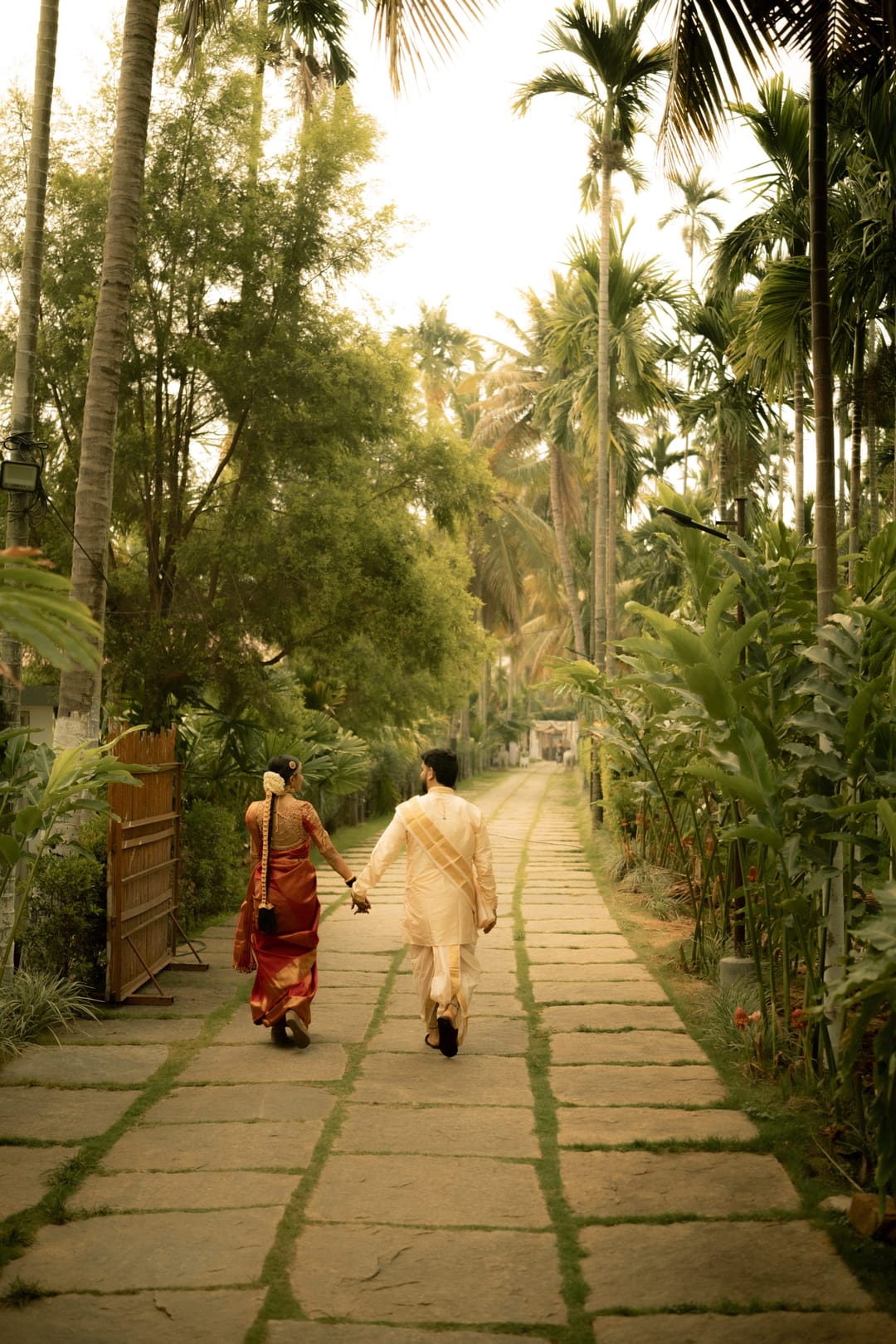 A couple in traditional clothes walks hand in hand down a stone path lined with areca palms.