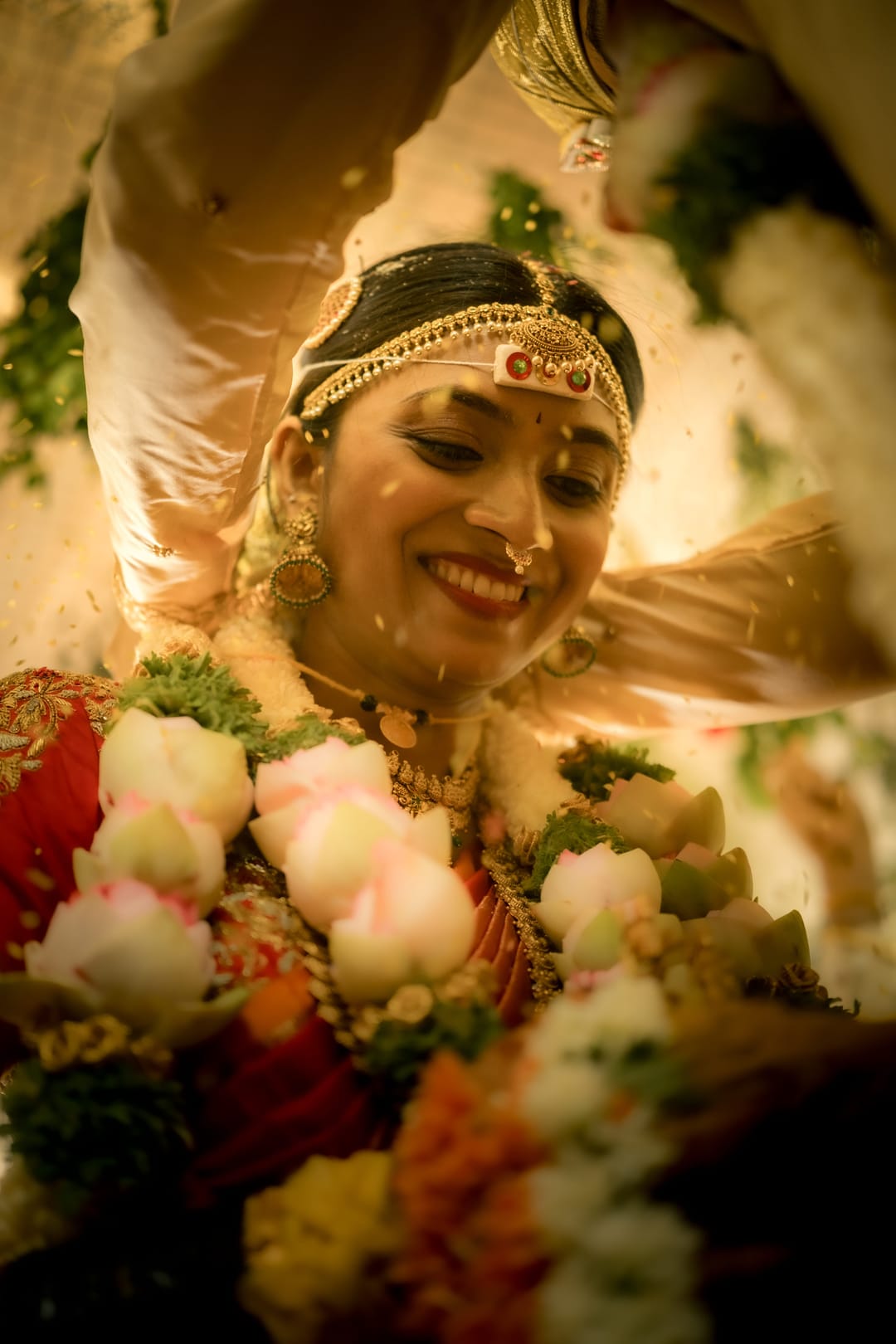 Framed by her garland of roses and jasmine, the bride smiles up during the ceremony in warm light.