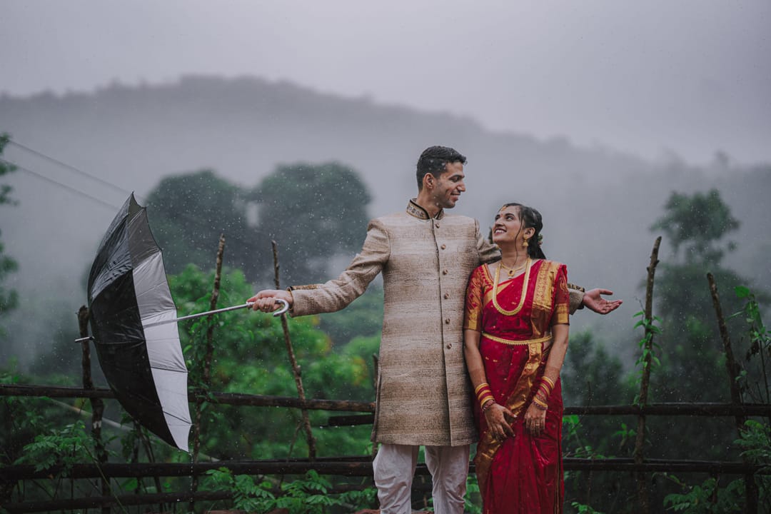 On a misty hilltop, the groom in a sherwani holds out a black umbrella beside the bride in a red saree.