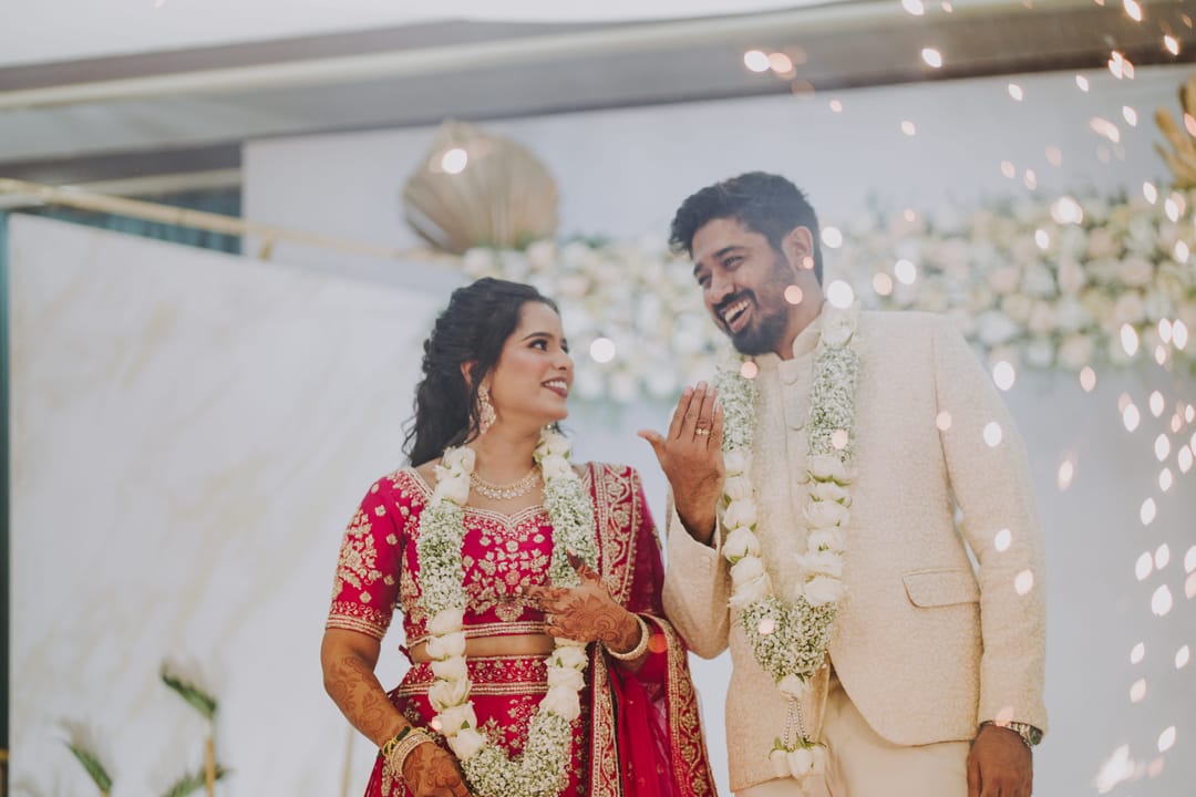 The bride and groom laugh together as petals fall around them, both wearing jasmine garlands.