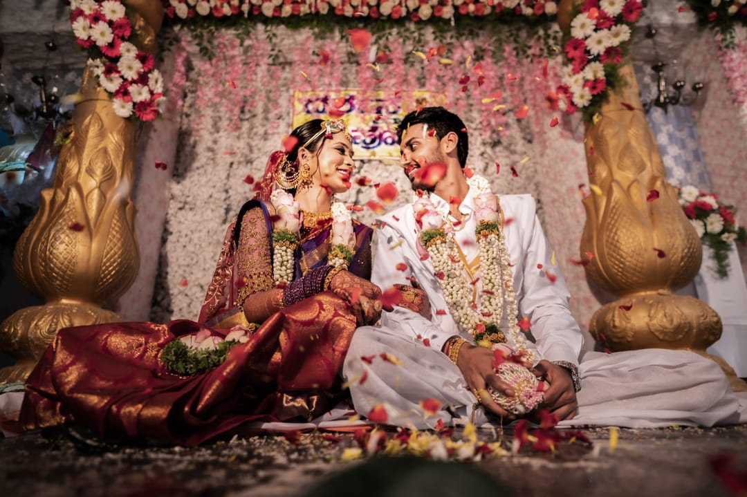 The bride and groom sit on a flower-strewn mandap, laughing as petals fall.