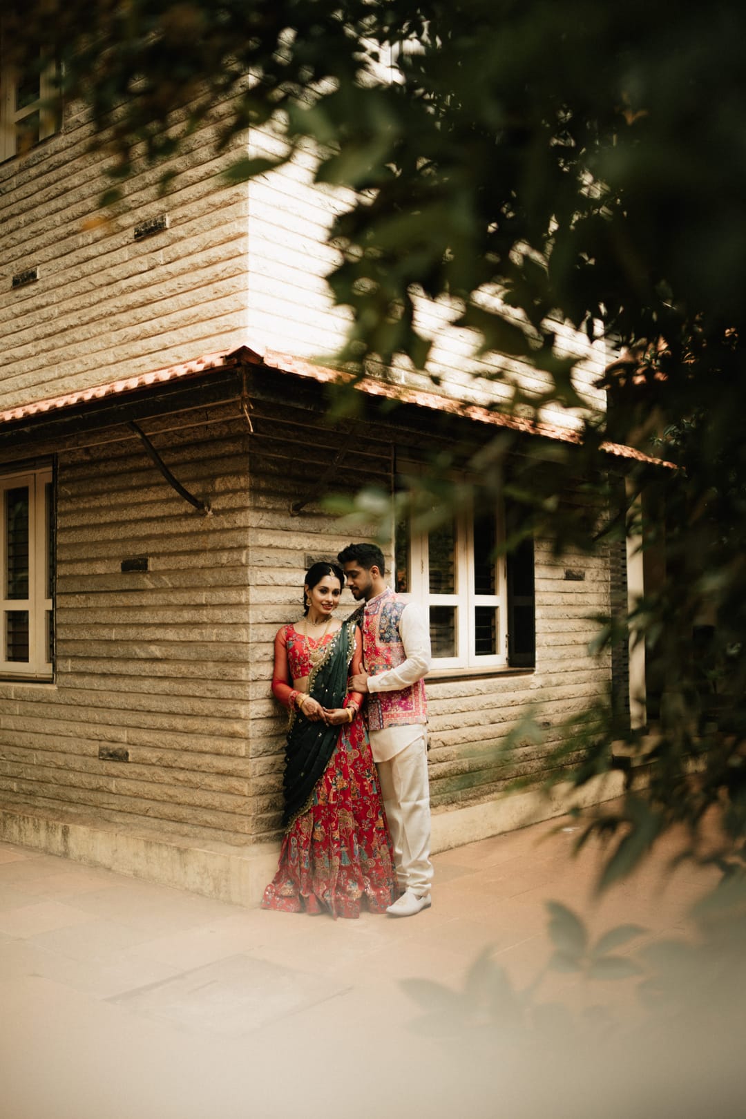 A couple leans against a log-walled cottage, framed by leaves, she in a red lehenga.