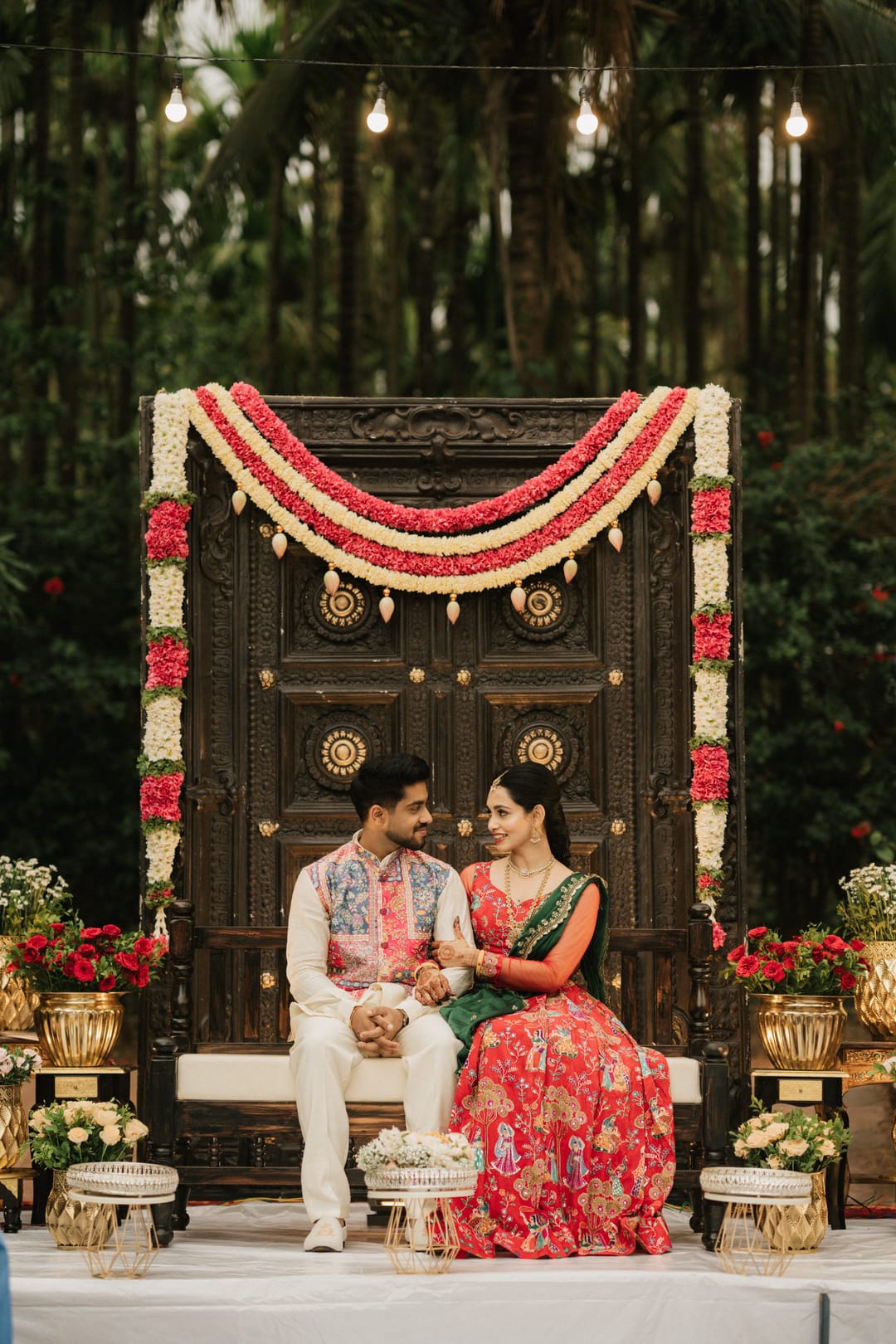 A couple sits on a carved wooden stage under marigold and rose garlands, laughing together.