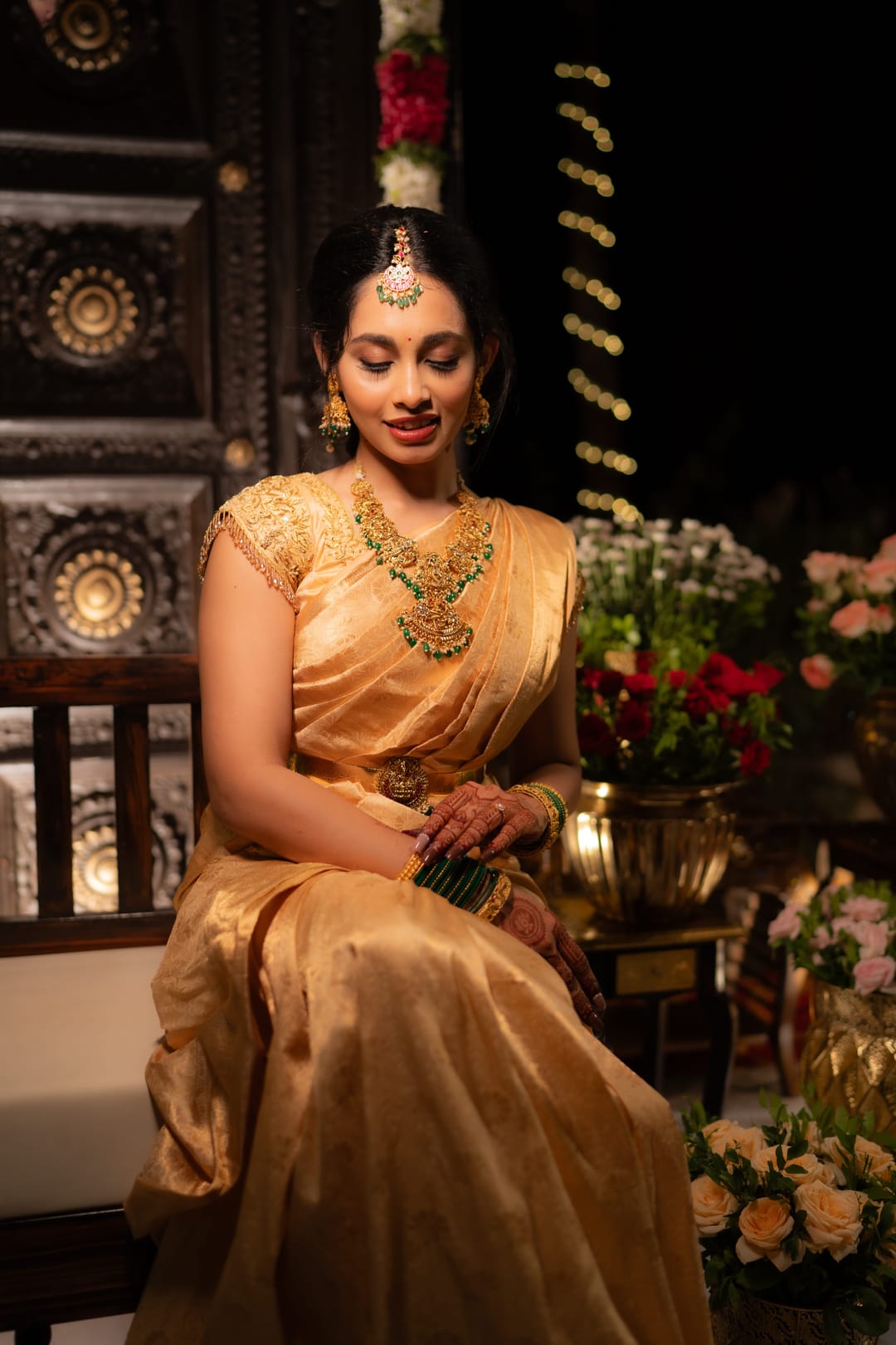 A bride in a gold silk saree sits among flowers before a carved dark wood backdrop, eyes lowered.