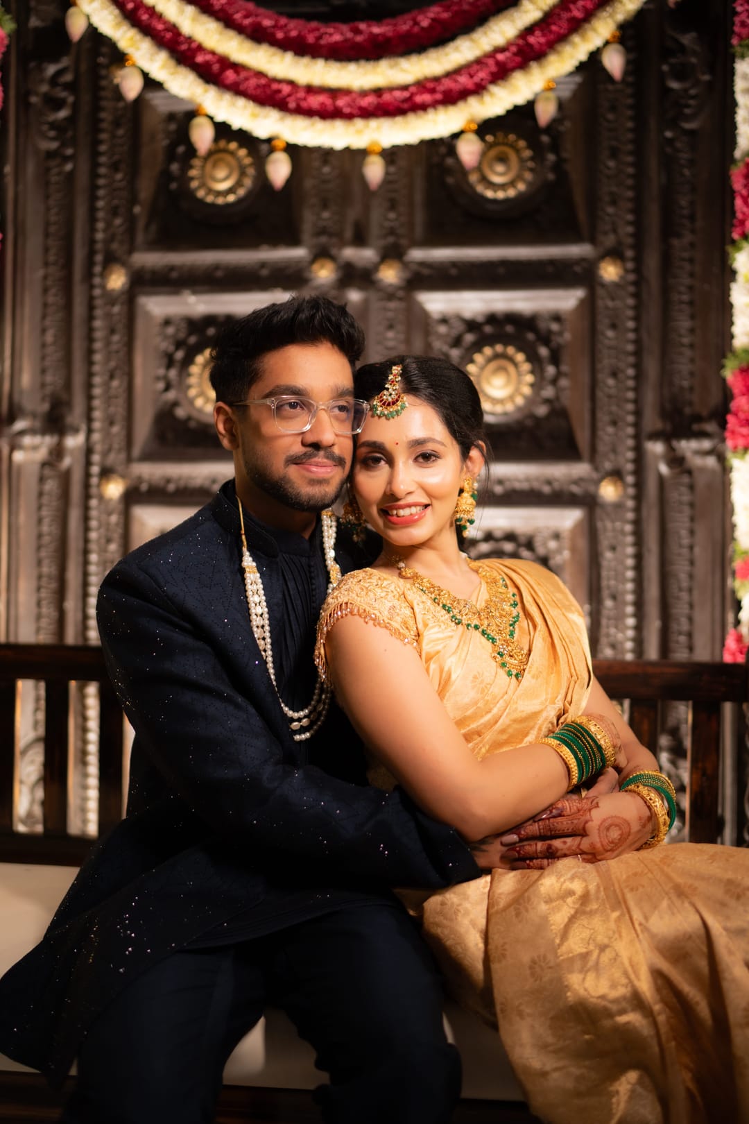 The bride and groom sit close in front of a carved door, smiling at the camera.