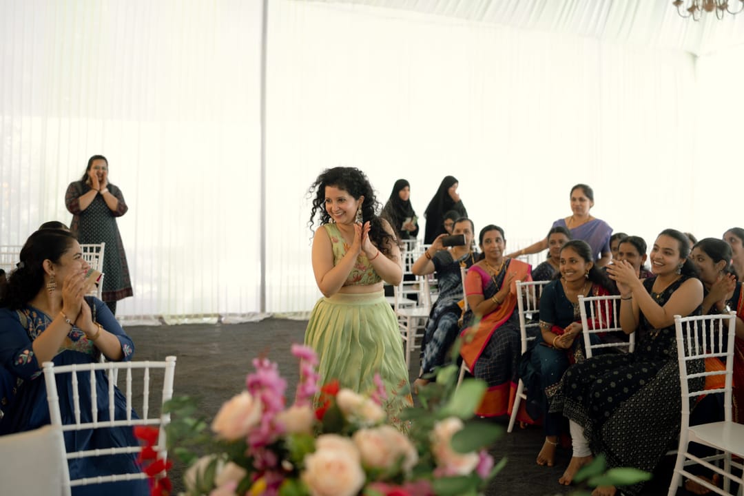 Guests clap and cheer as a woman in a green lehenga dances down the aisle of a white marquee.