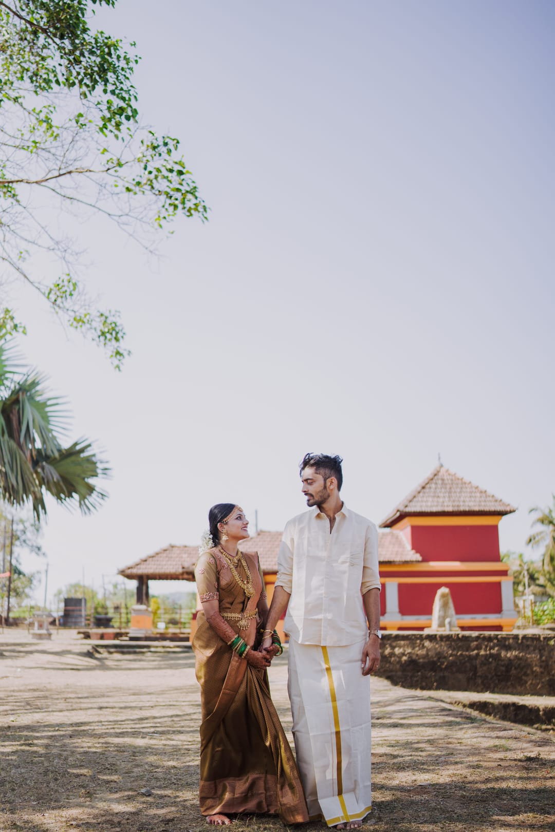 A couple stands holding hands in front of a red-roofed temple under a pale sky.