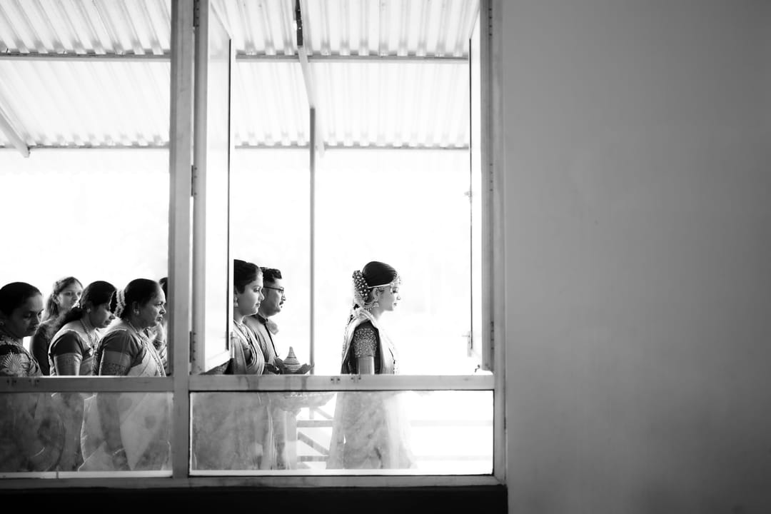 Black-and-white: women in saris pass a row of tall windows, silhouetted against the light.