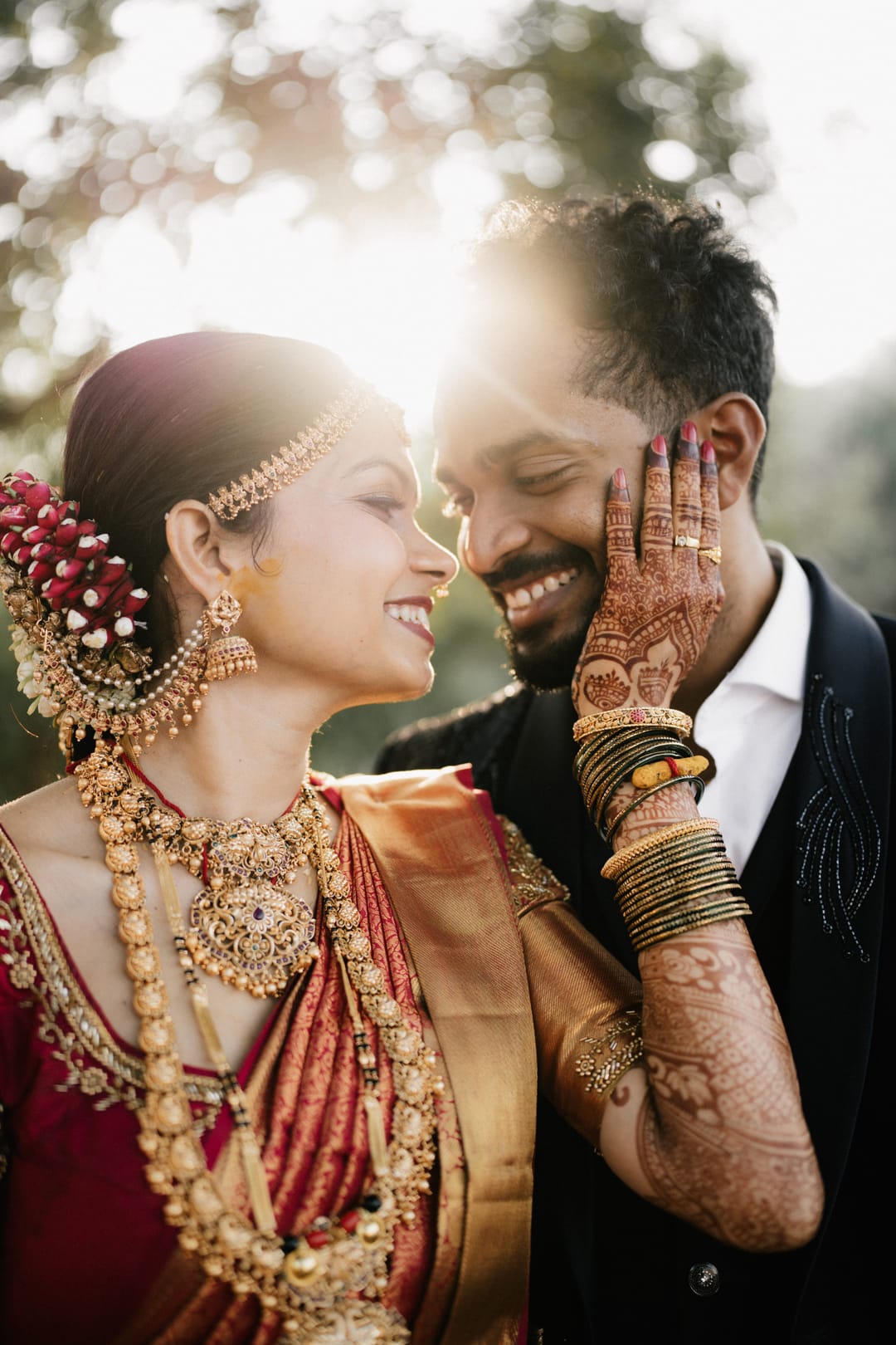 In soft backlight, the groom cups the bride's face as she smiles, her hair adorned with roses.