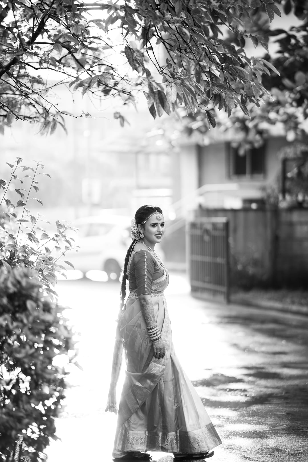 Black-and-white: a bride in a long skirt walks down a wet street framed by overhanging leaves.