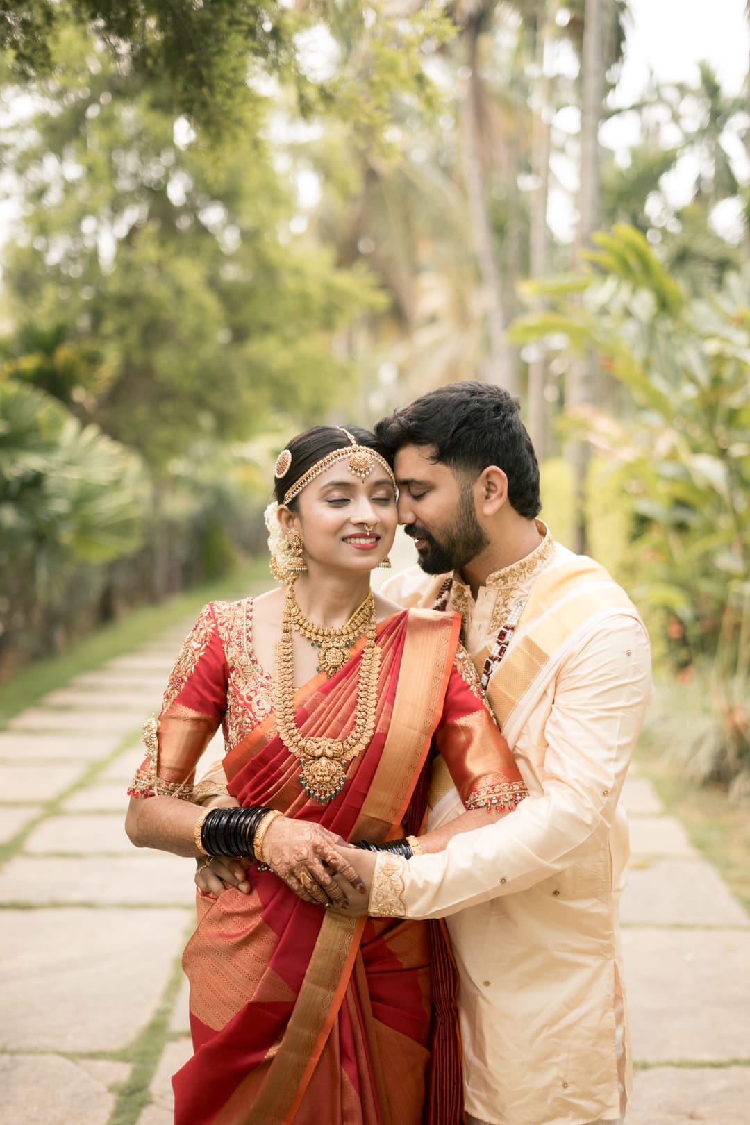 On a garden path, the groom holds the bride from behind as she smiles, eyes closed.