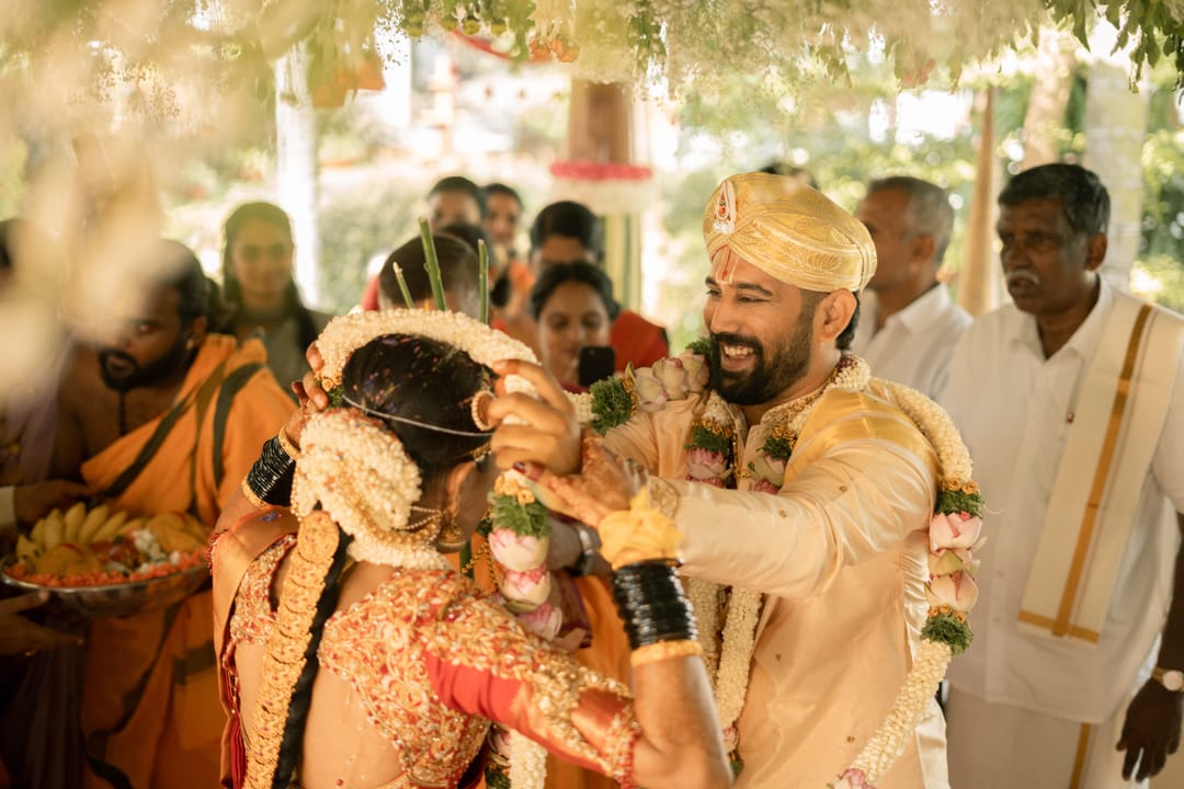 The groom places a garland over the bride's head as priests and family look on.