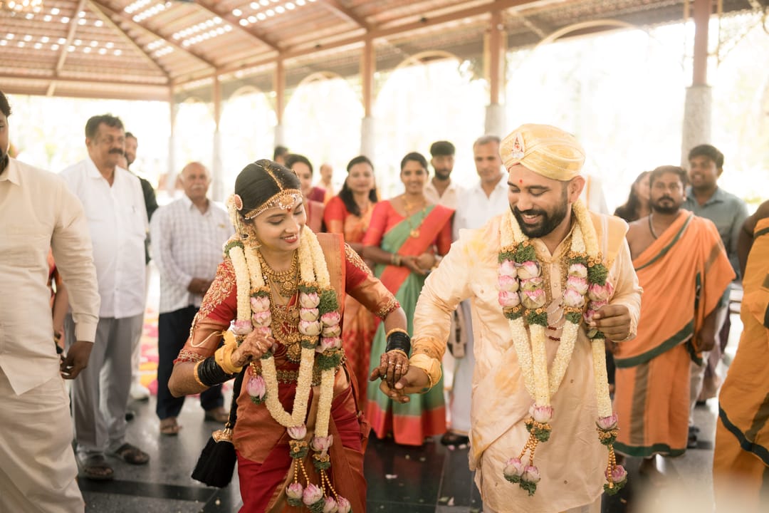 The bride and groom, garlanded, walk hand in hand through a pavilion full of guests.