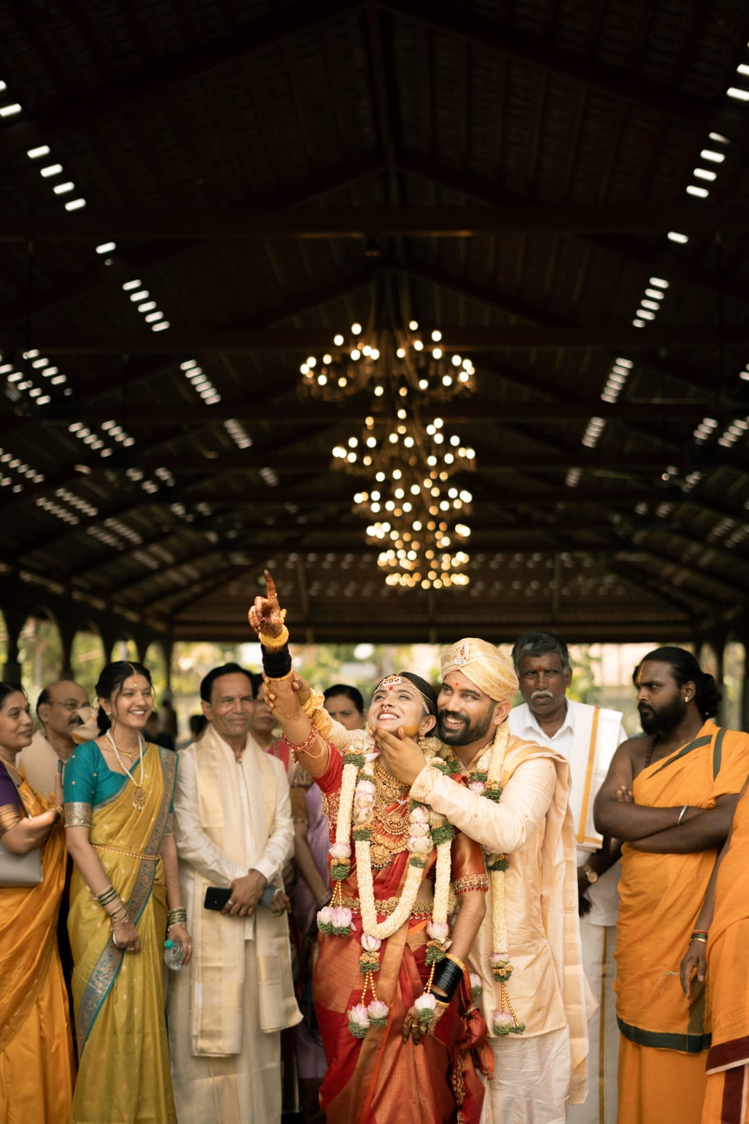 Under chandeliers in a timber pavilion, the bride points up and laughs as the groom holds her, guests smiling around them.