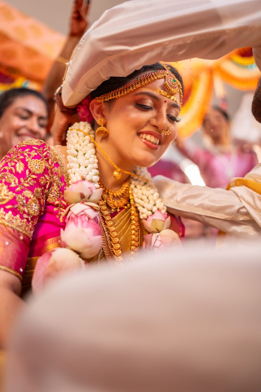The bride beams beneath a white cloth canopy, roses and jasmine garlanded around her neck.