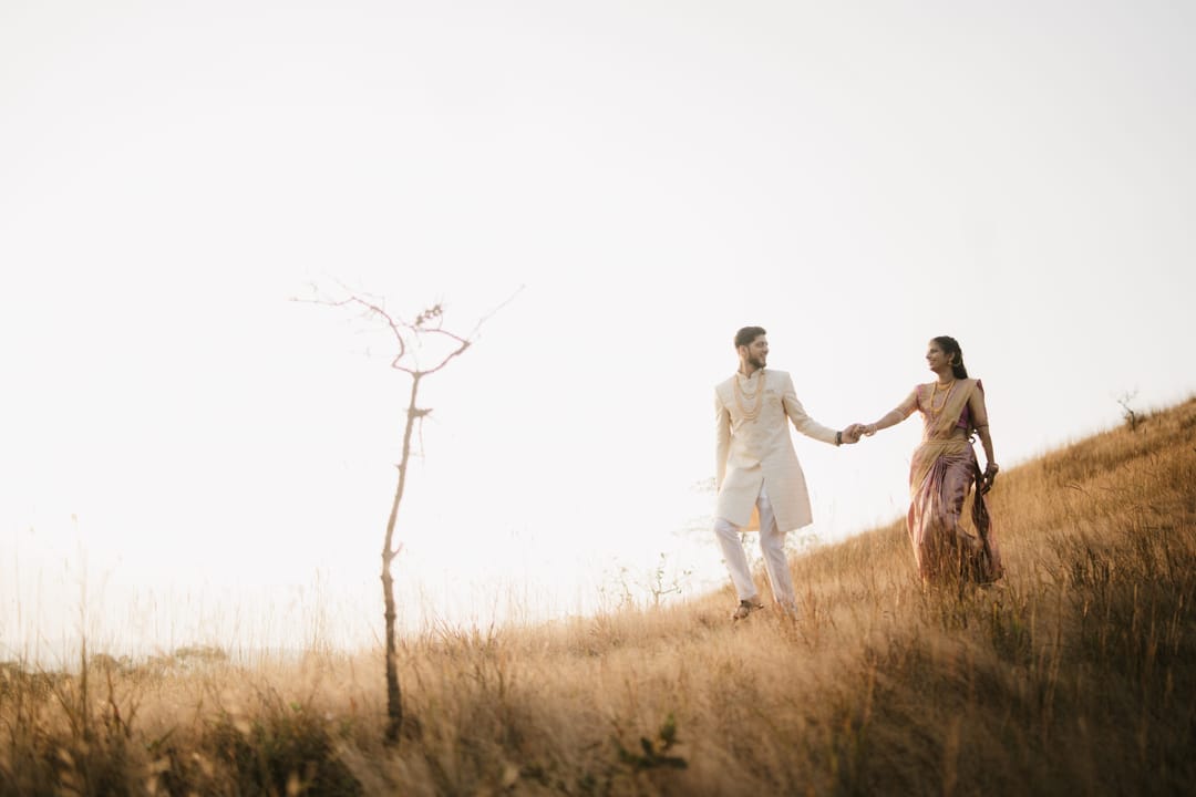 Far off on a dry golden hillside, a couple walks hand in hand past a lone bare tree.