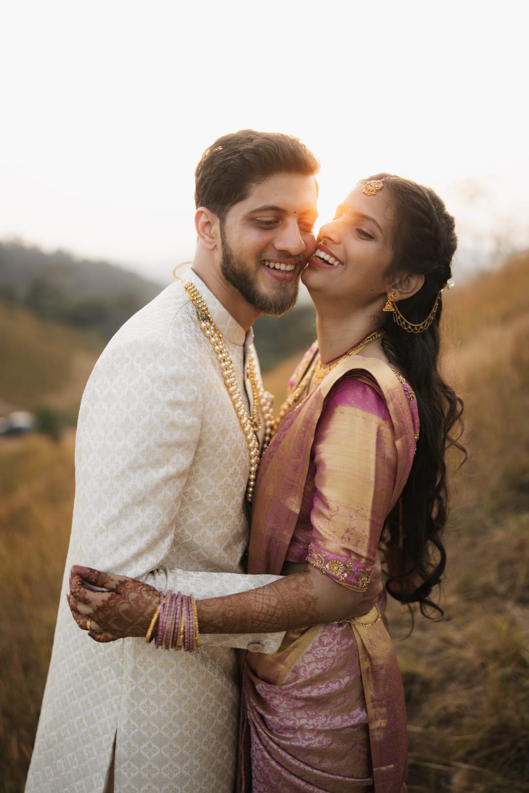 A couple laughs in an embrace on a hillside at sunset, she in a pink silk saree.