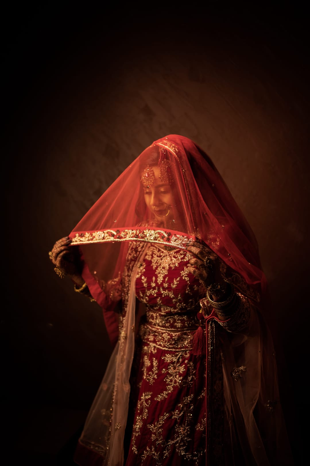 A bride under a red embroidered veil looks down, lit softly against a dark background.