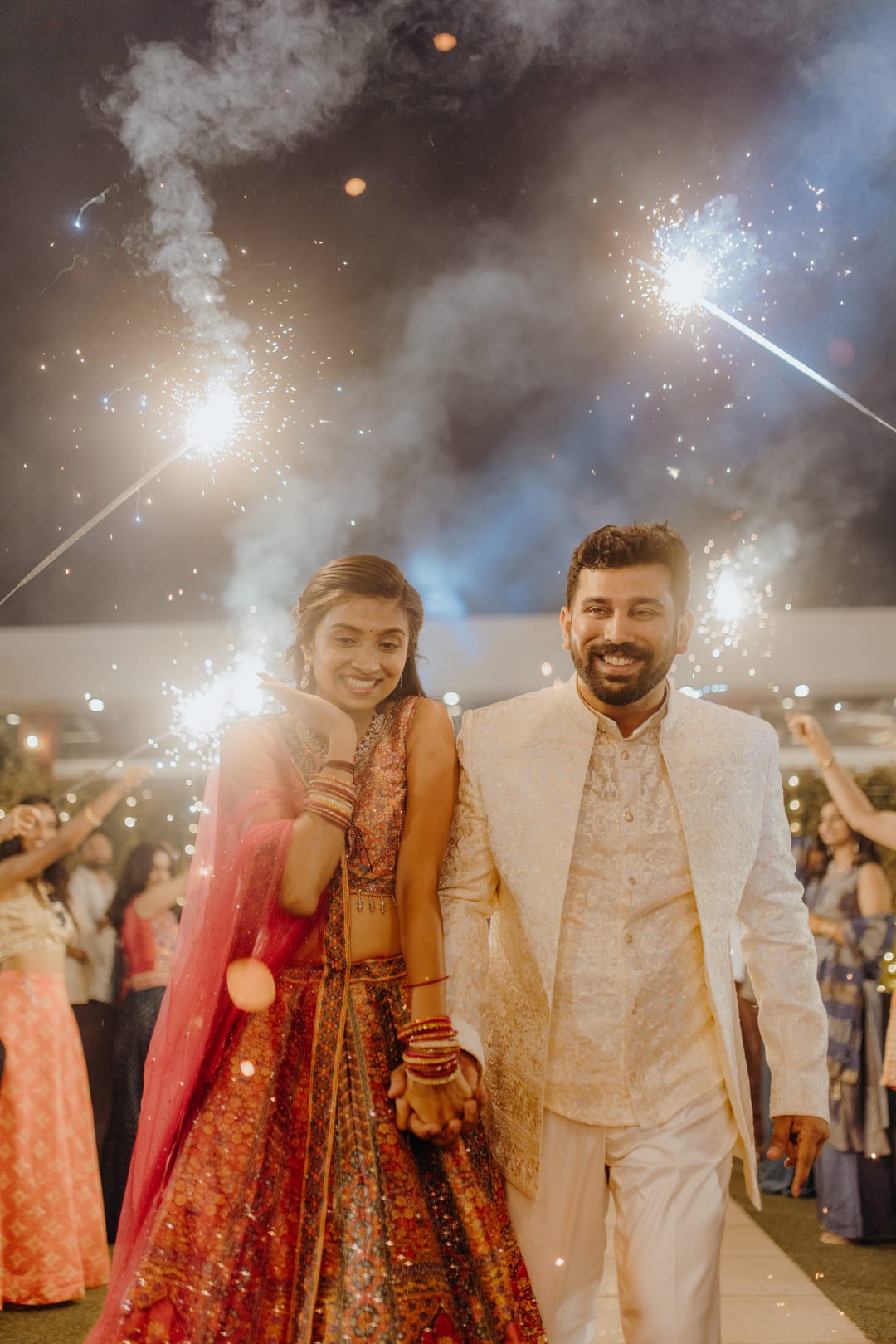 The couple walks through a shower of sparklers, she in a red lehenga, he in an ivory sherwani.